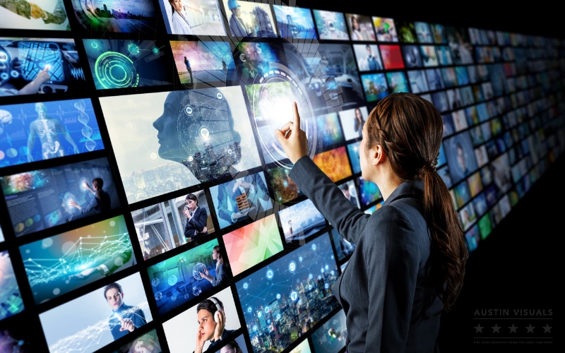 A businesswoman interacts with a large digital video wall displaying multiple futuristic screens showing technology, data, people, and innovation visuals, representing advanced video marketing and digital communication concepts. Austin Visuals logo appears in the lower right corner
