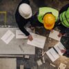 Overhead view of four workers in safety helmets and high visibility vests gathered around a table with blueprints and tools in an industrial setting