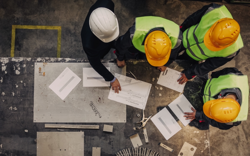 Overhead view of four workers in safety helmets and high visibility vests gathered around a table with blueprints and tools in an industrial setting