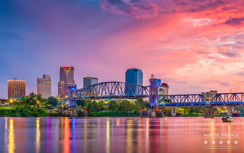 View of downtown Little Rock Arkansas featuring the illuminated bridge reflecting on the calm river under a vibrant sunset sky Austin Visuals logo displayed