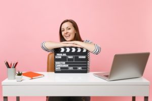 Young laughing woman leaning on classic black film making clapperboard and working on project while sitting at office with laptop